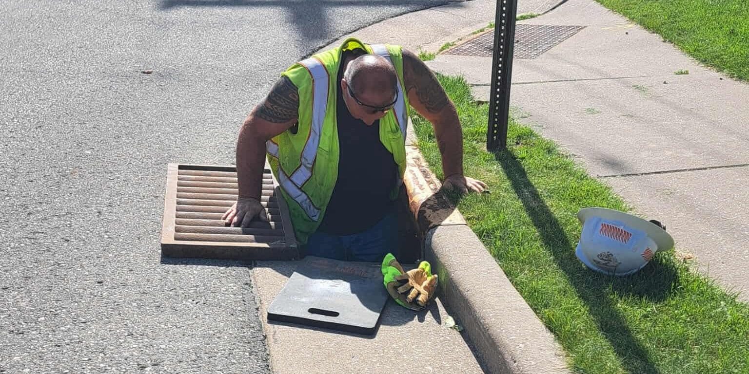 A We Energies employee lowers himself into a storm sewer in a roadway next to a sidewalk.