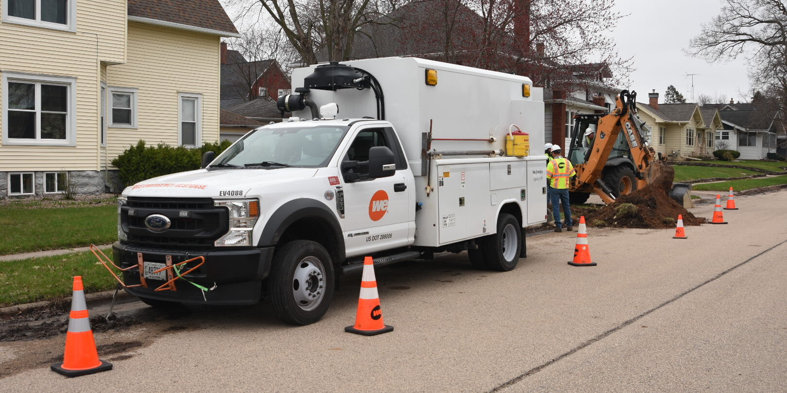 A large We Energies truck is parked along a street with an excavator parked behind the truck.